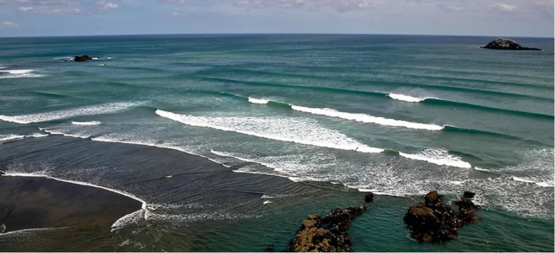 A clean SW groundswell lining up across the bay. Multiple peaks running through, the southern bank visible in the far right, 